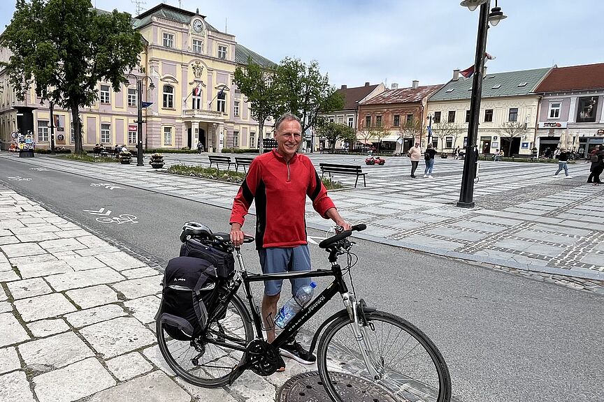Zeitreise Liptovsky Mikulas Ein Mann hält ein Fahrrad vor einem Radweg. Im Hintergrund ist ein Platz zu sehen, an dem ein Rathaus steht.